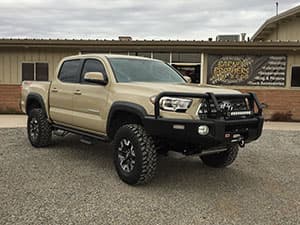 A beige, lifted Toyota truck is parked on gravel in front of a matching beige building under a cloudy sky.