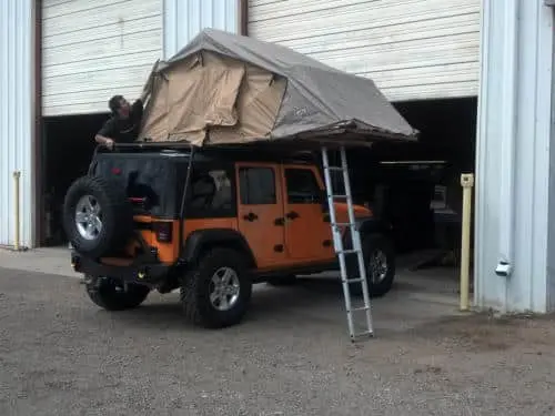 An orange jeep with a rooftop tent and attached ladder, part of our impressive customer rigs, is parked in front of a garage where a person is diligently working on the tent.