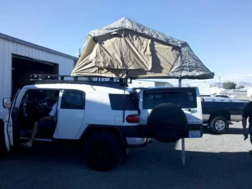 A white SUV with a rooftop tent and a black truck, both examples of impressive customer rigs, parked beside a building in a sunny outdoor area.
