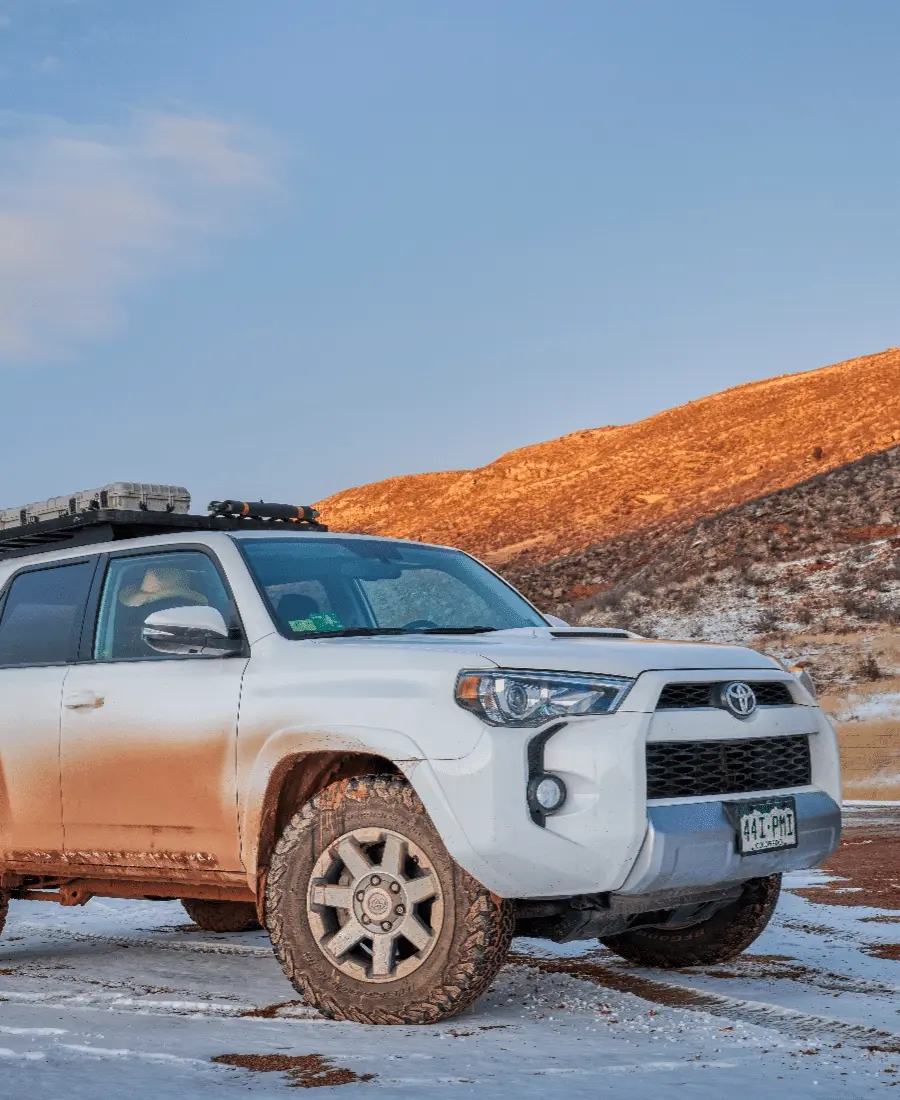A white SUV parked on a snowy dirt road near Grand Junction, with the sunrise illuminating the rocky hills in the background. The scene looks like something straight out of a Barney Brothers advertisement, capturing both rugged beauty and serene tranquility.