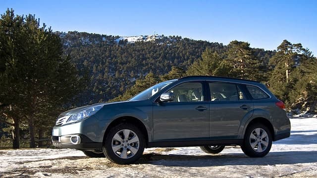 A blue Subaru station wagon is parked on a snowy landscape, with majestic mountains and trees in the background.