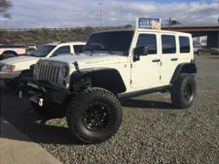 A white Jeep Wrangler with oversized tires, one of the impressive customer rigs, parked on a gravel surface, with another vehicle in the background.