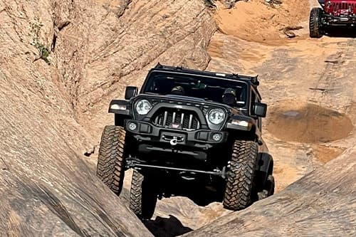 A rugged off-road vehicle with impressive 4x4 modifications traverses rocky terrain, delivering off-road thrills, with another vehicle in the background.