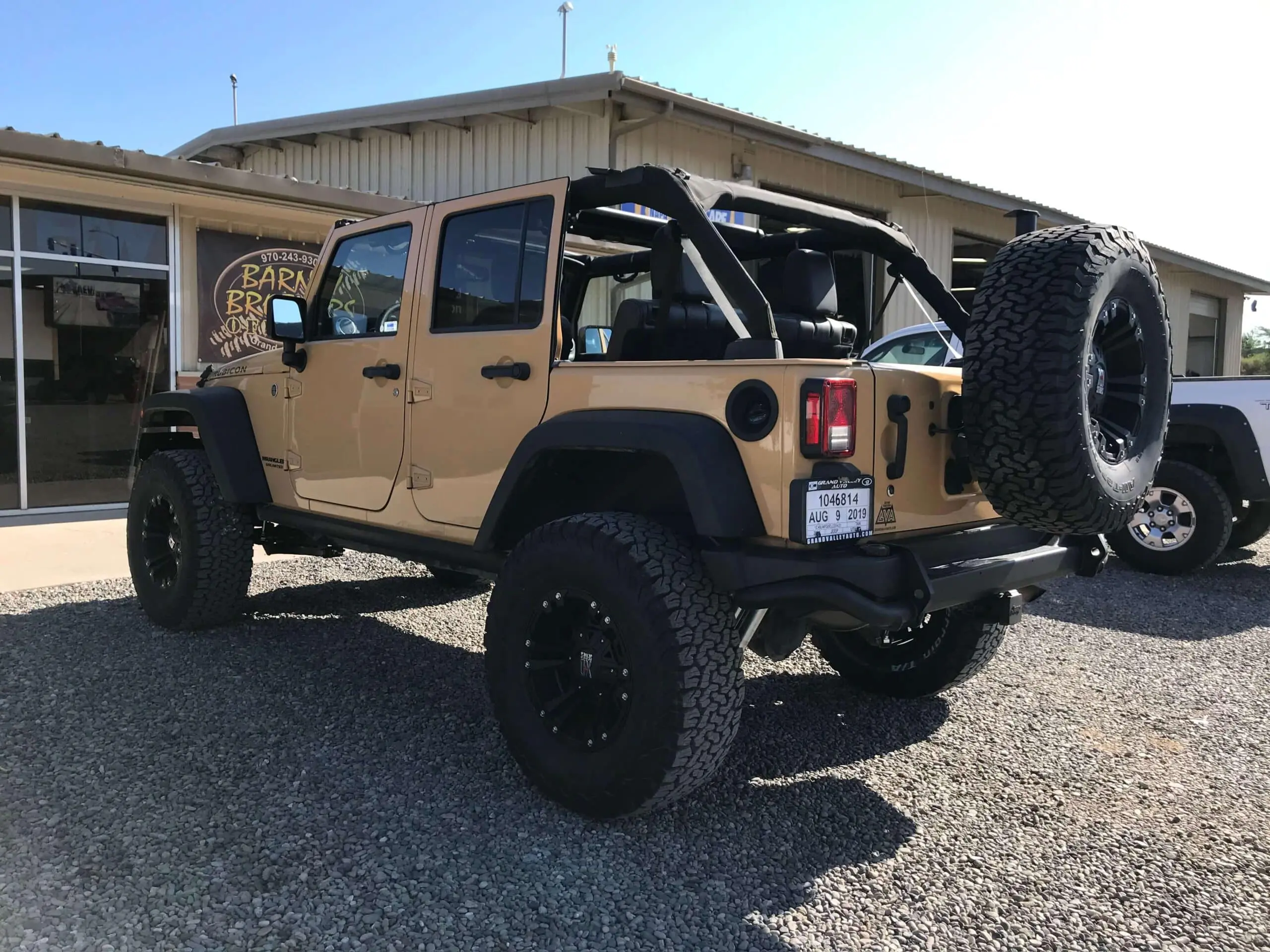 A tan, four-door Jeep Wrangler with off-road mod tires is parked outside a building with a sign that reads "Barn.