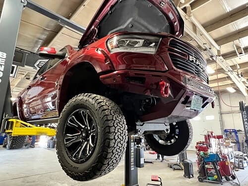 A red truck with off-road tires is lifted in a garage for auto repair and maintenance. Tools and equipment are visible around.