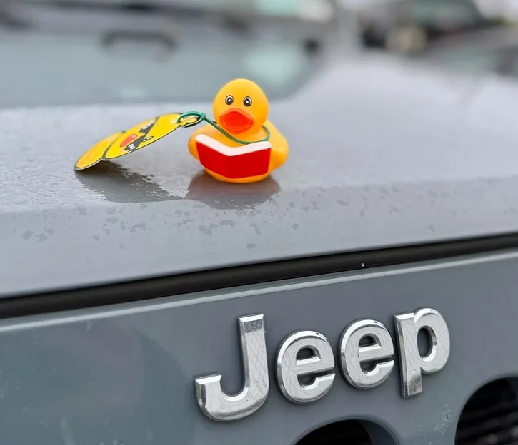 Blue rubber duck and pink pixel-style duck displayed on the hood of a Jeep in front of a round headlight, representing the Jeep Ducking community tradition.
