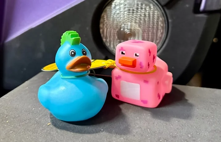 Blue rubber duck and pink pixel-style duck displayed on the hood of a Jeep in front of a round headlight, representing the Jeep Ducking community tradition.