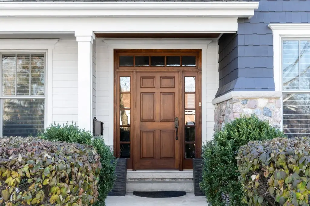 A front door and covered porch detail on a home with grey siding, white trim, red brick, a wooden front door.
