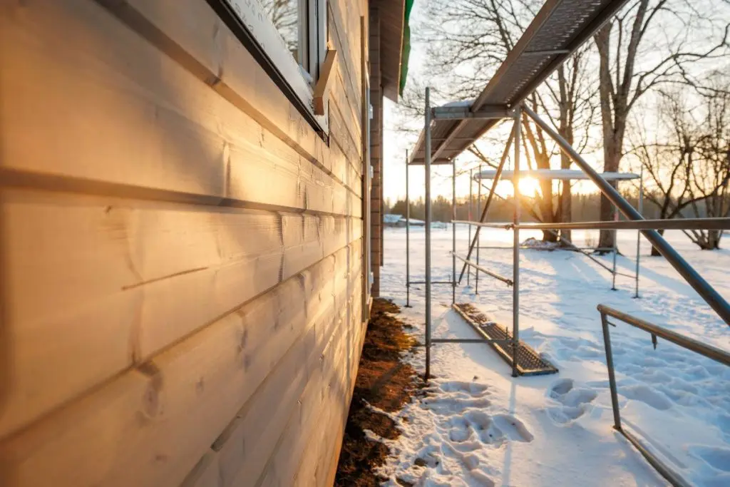 Close up wooden siding glows in warm late afternoon light as metal scaffolding lines a narrow snowy path with footprints at a rural residential build