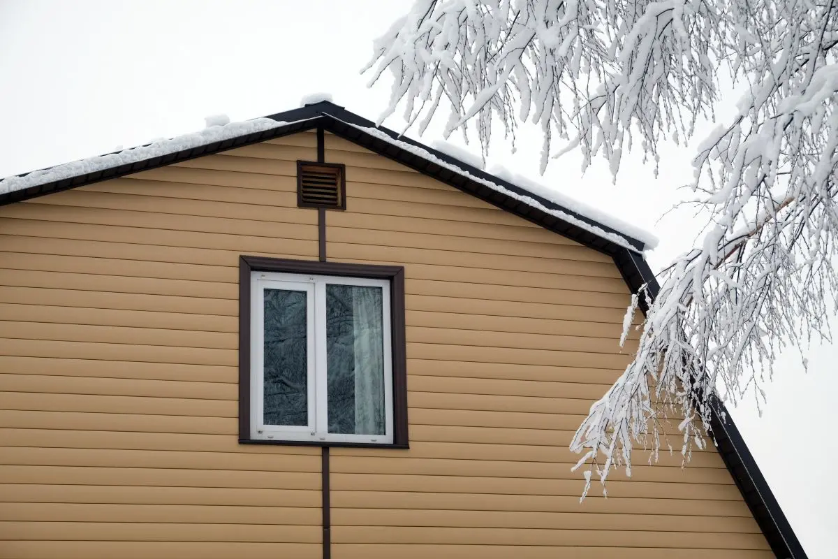 Upper floor with plastic window in country house wall covered with beige siding and brown metal roof with snow