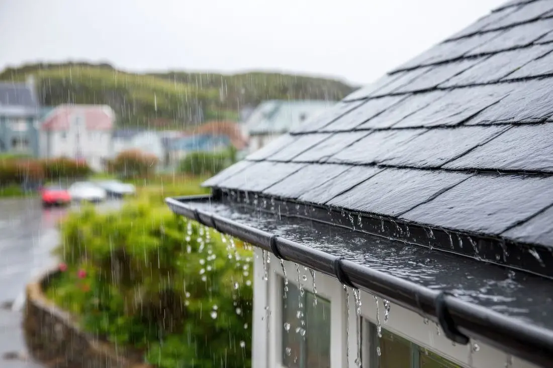Rainwater flows down a roof and gutter during a downpour, blurring the background with heavy rainfall.