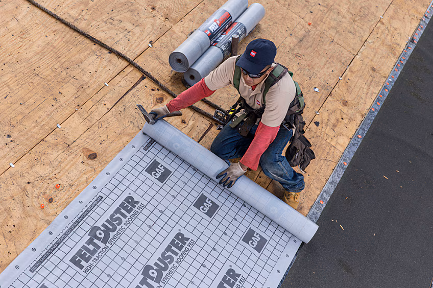 This shows a laborer installing a roofing system by GAF and they are rolling out felt paper which is one of the variables that will decide how much your roof costs