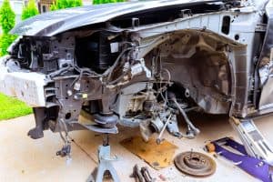 Front view of a car missing its bumper and front wheel, showing exposed mechanical parts and wiring. The car is propped on a jack stand, conveying a tone of repair or disassembly.