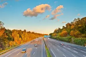 A highway under a clear blue sky with fluffy clouds; surrounded by autumn-colored trees. Cars are moving smoothly, conveying a calm, serene atmosphere.