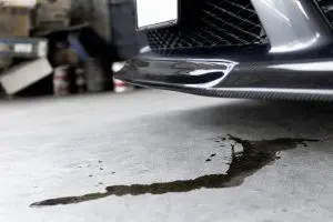 Close-up of a car's front bumper with a puddle of oil on a garage floor, suggesting a leak. The scene conveys maintenance needs, with a calm atmosphere.