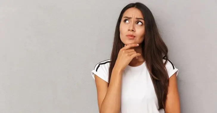 woman thinking while looking up and holding hand to chin against a gray wall