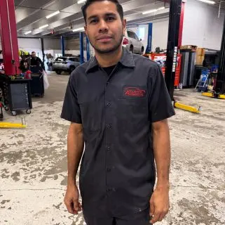 A man in a dark work uniform stands in an automotive repair shop, surrounded by lifts, tools, and cars. As one of the auto repair experts in Northglenn, he looks at the camera with a neutral expression.