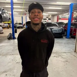 A man wearing a black hoodie and a black cap stands smiling in an auto repair shop, with cars and equipment in the background, showcasing the friendly team of auto repair experts in Northglenn.