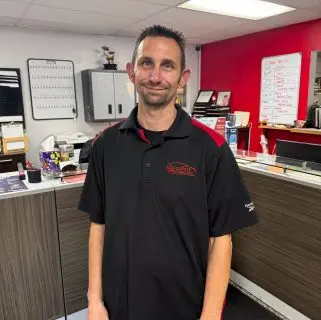 A man wearing a black polo shirt with a red logo stands inside an office with a counter, paperwork, and various items in the background. He is smiling and has short dark hair. The office of these auto repair experts in Northglenn has red and white walls.