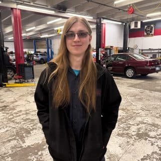 A person with long blond hair, glasses, and a black jacket stands smiling inside an auto repair shop—home to auto repair experts in Northglenn—with cars, lifts, and equipment visible in the background.