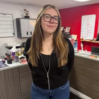 A young woman with long, straight hair and glasses stands indoors, wearing a black zip-up hoodie and blue jeans. She is in an office with a red and white wall, where auto repair experts in Northglenn manage various tasks at their desks.