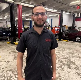 A man wearing glasses and a black work shirt with a red logo stands smiling in an auto repair shop, embodying the trusted auto repair experts in Northglenn, surrounded by cars and vehicle lifts.