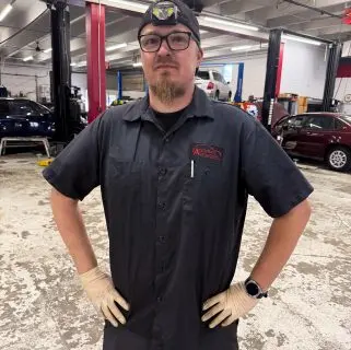 A mechanic, one of the auto repair experts in Northglenn, wearing gloves, glasses, and a black work shirt stands confidently in an auto repair shop with cars and equipment in the background.