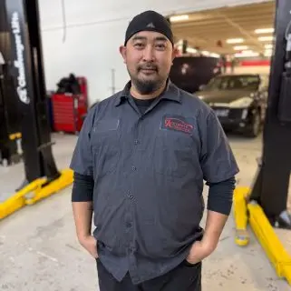 A man wearing a dark button-up work shirt and a black head covering stands in an auto repair shop with cars, lifts, and tools—typical of auto repair experts in Northglenn—visible in the background.