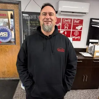 A man with short dark hair and a gray goatee, wearing a black hoodie, stands smiling with his hands in his pockets inside an office of auto repair experts in Northglenn, with motivational posters on the wall behind him.