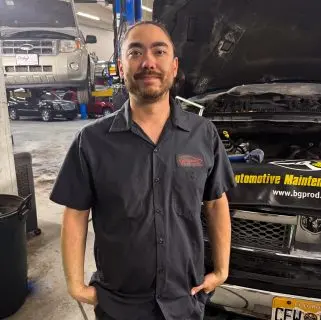 A mechanic wearing a dark uniform stands in a garage in front of a car with its hood open, showcasing the expertise of auto repair experts in Northglenn. Another vehicle is raised on a lift, surrounded by tools and equipment.