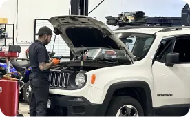 A mechanic wearing gloves works under the raised hood of a white Jeep SUV inside an auto repair shop, using a diagnostic tool. Various tools and equipment are visible in the background.