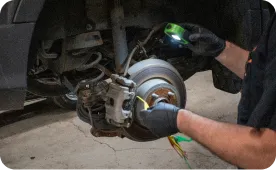 A mechanic wearing black gloves inspects a car’s exposed brake system with a flashlight, checking the brake rotor and caliper. The car is elevated, and tools are visible in the mechanic’s hands.