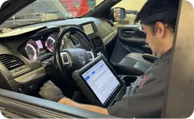 A mechanic sits in a cars driver seat, using a diagnostic tablet connected to the vehicles system. The dashboard displays lights, and the technician wears black gloves, working in a garage setting.