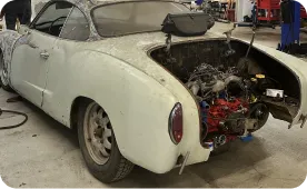A dusty vintage white car with its rear hood open, exposing the engine and internal mechanical parts, is parked inside a workshop or garage.
