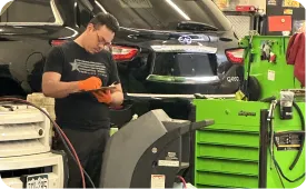 A mechanic wearing orange gloves writes on a clipboard in an auto repair shop, standing near a black SUV and green tool chest, with various equipment and tools around him.