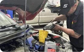 A mechanic wearing a cap pours engine oil into a yellow funnel under the hood of a car in a garage, with oil containers and tools visible nearby.