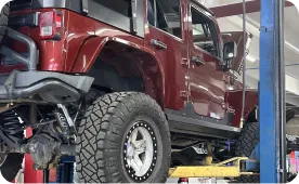 A red Jeep Wrangler with large off-road tires is elevated on a hydraulic lift inside an auto repair shop.