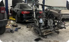 A black car is parked in an auto repair shop with its hood open. An engine has been removed and is suspended by a hoist nearby. Tools and car parts are scattered on the dirty shop floor.