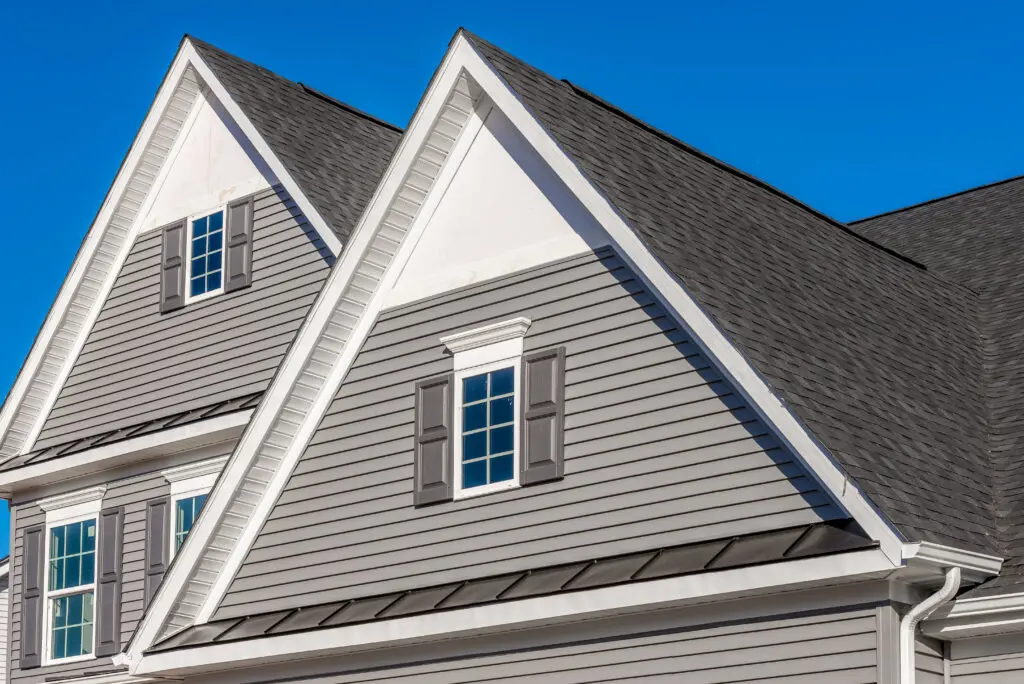 Double gable, with white decorative trim over the windows on a triangle gable roof, white soffit and fascia, gray  horizontal vinyl lap siding with blue sky background fascia board replacement