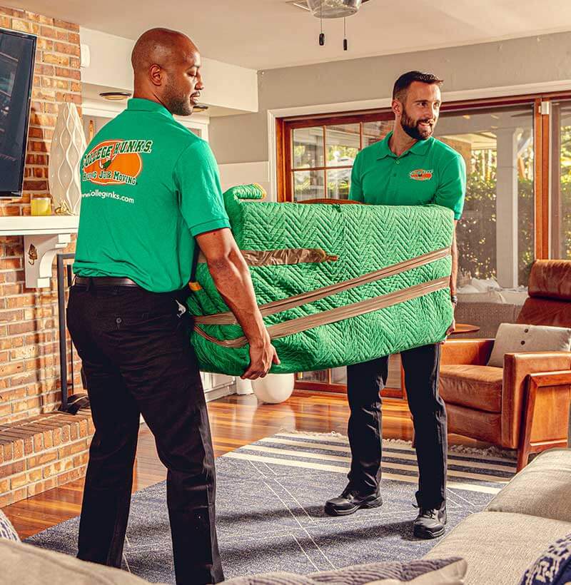 Two uniformed College HUNKS movers carrying a padded piece of furniture through a living room, the daily work a moving business plan prices