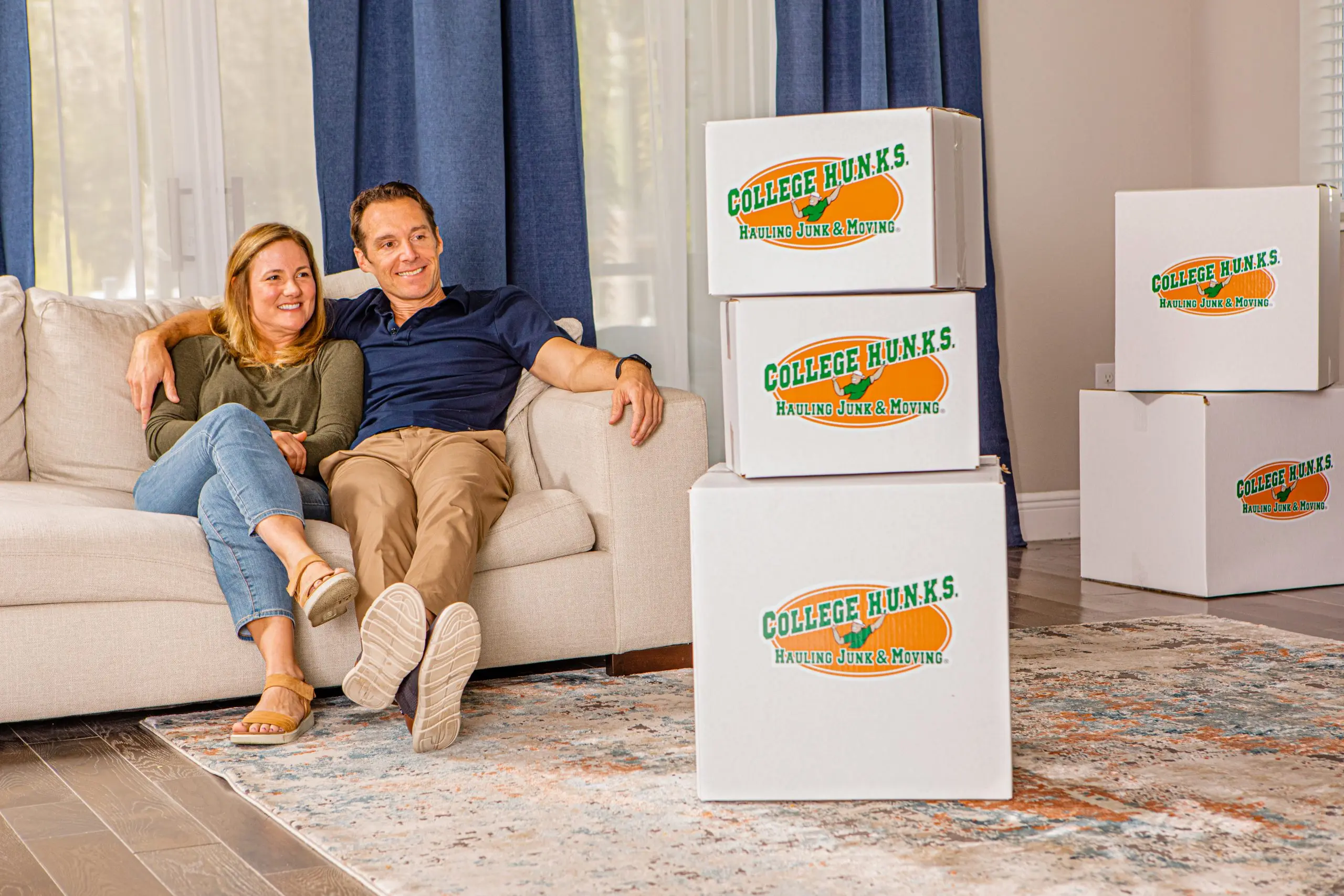 Couple relaxing on a couch beside College HUNKS moving boxes, planning the years between a layoff at 60 and Medicare