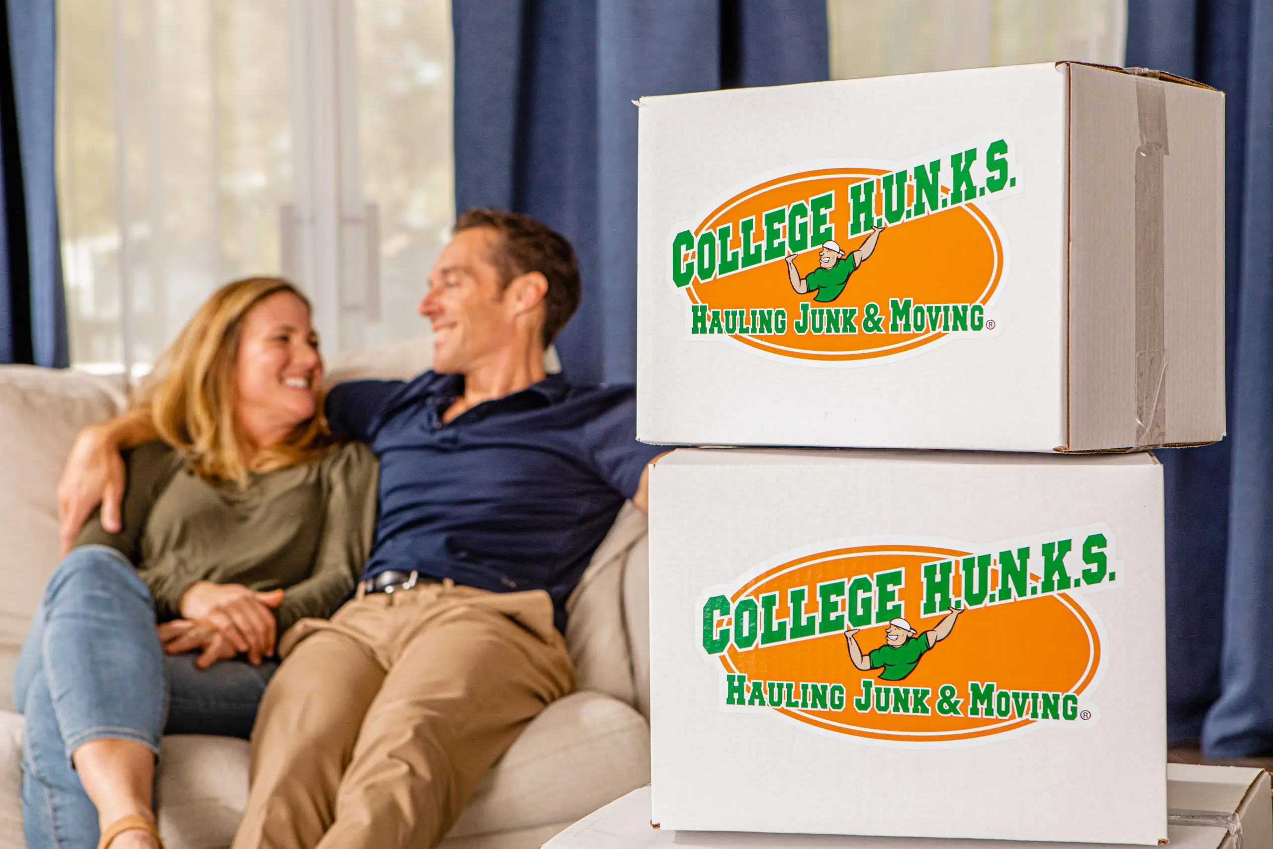 Couple relaxing on a couch next to stacked College HUNKS moving boxes