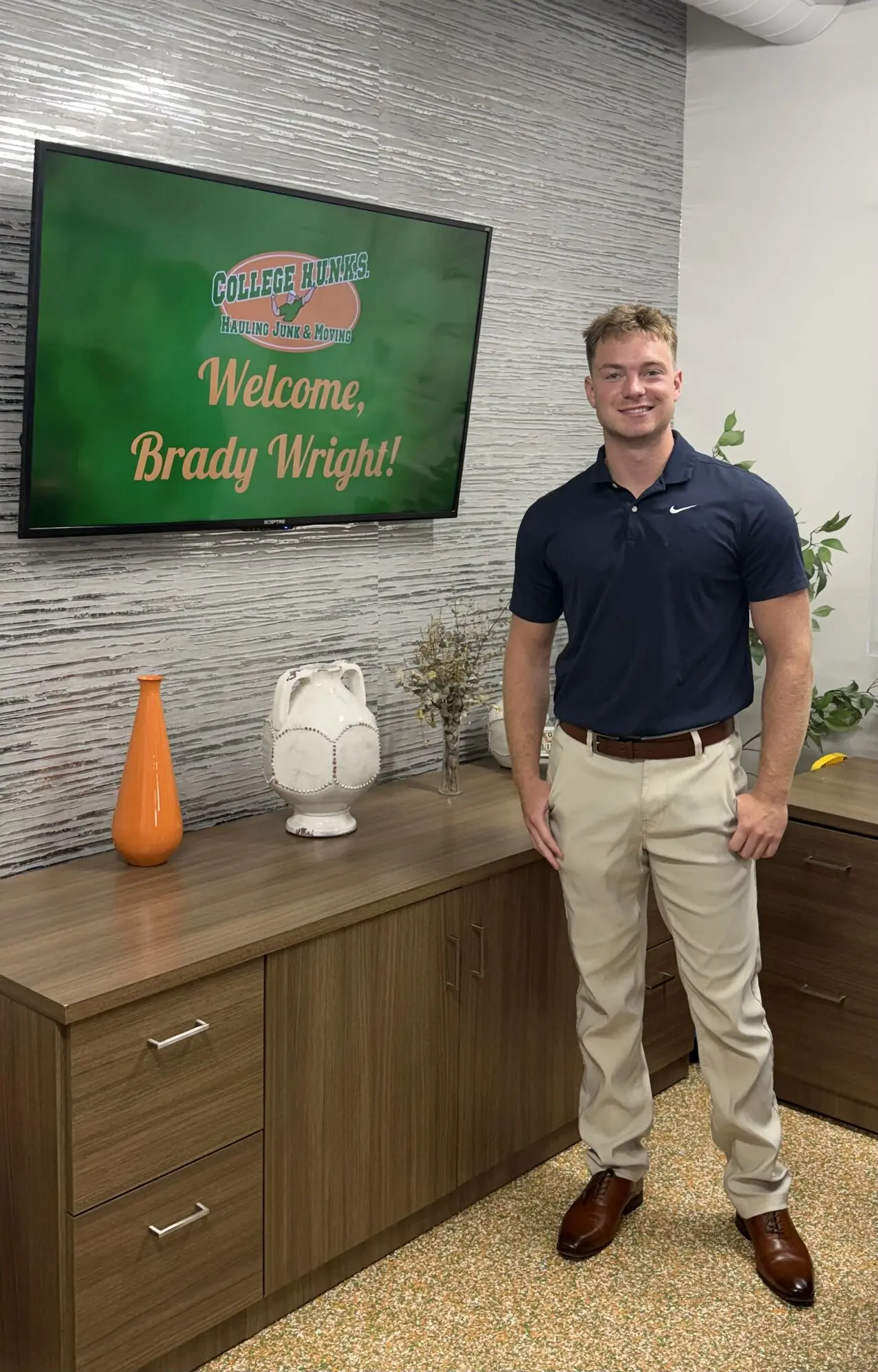 Brady Wright standing beside a Welcome, Brady Wright! screen at College HUNKS headquarters in Tampa, Florida