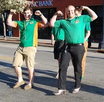 Two College HUNKS crew members in green polos flexing their arms on a city street