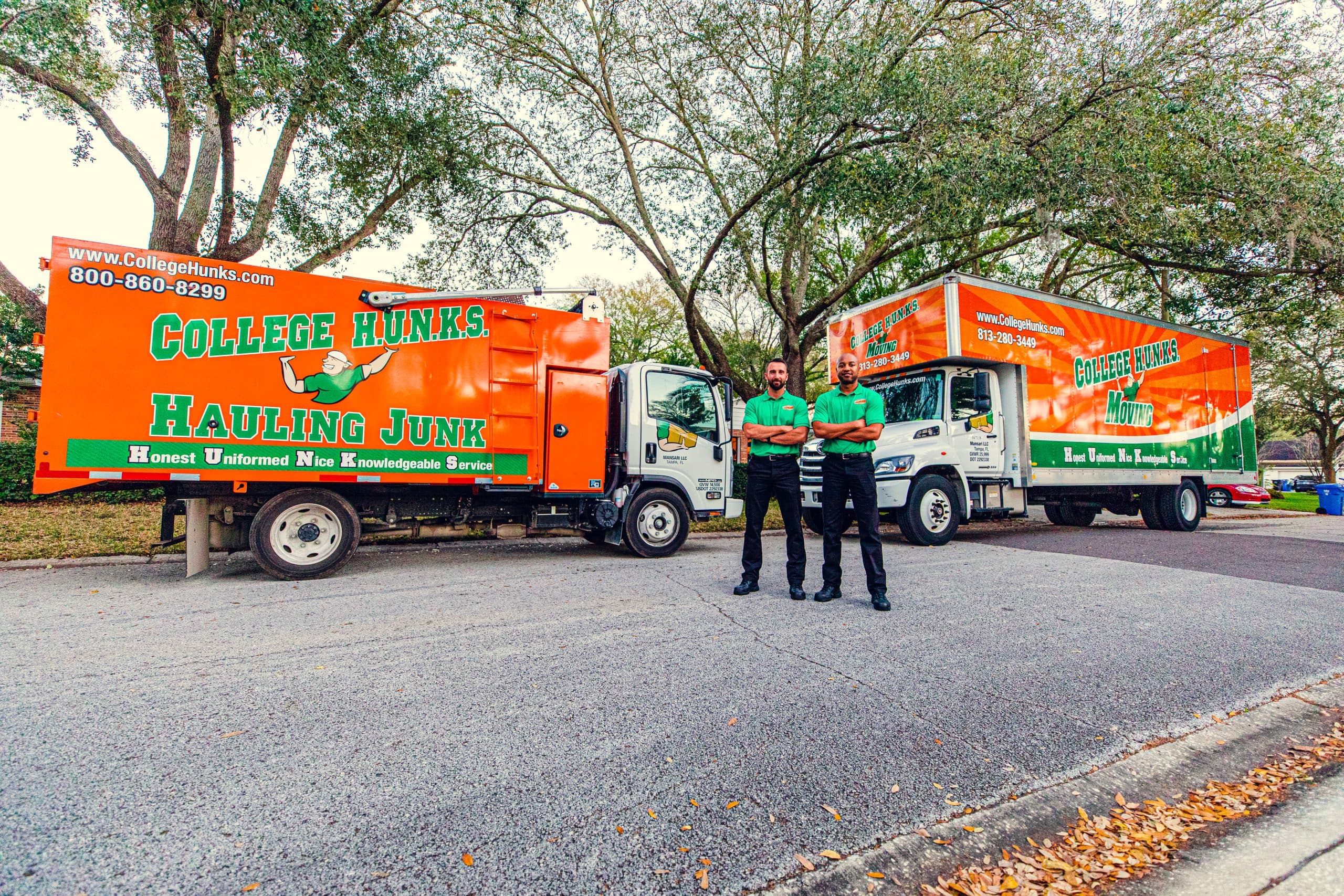 Two College HUNKS crew members standing between branded junk removal and moving trucks on a residential street