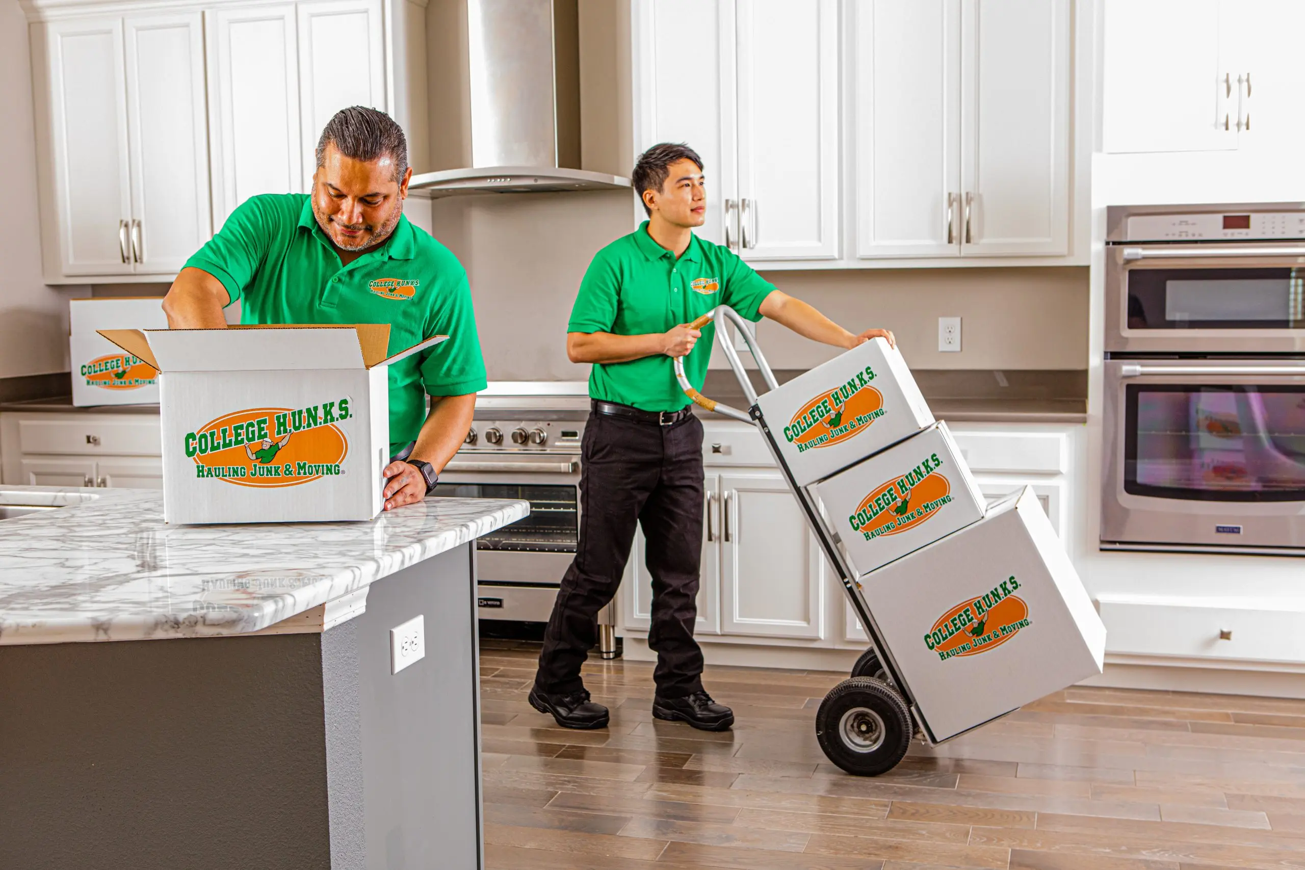 Two College HUNKS movers packing branded boxes onto a hand truck in a kitchen, the job a moving company business plan prices