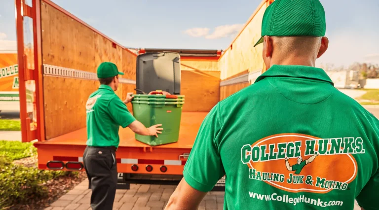 Two College HUNKS crew members in green uniforms loading storage bins into the back of an orange junk removal truck