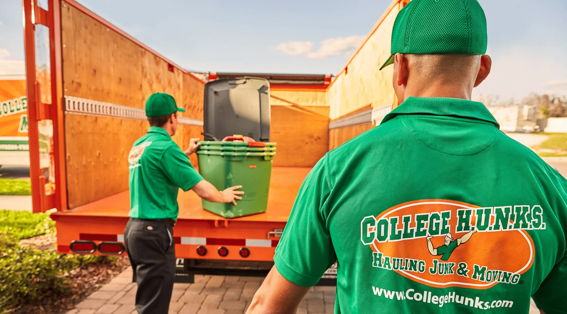 Two College HUNKS crew members in green uniforms loading storage bins into the back of an orange junk removal truck