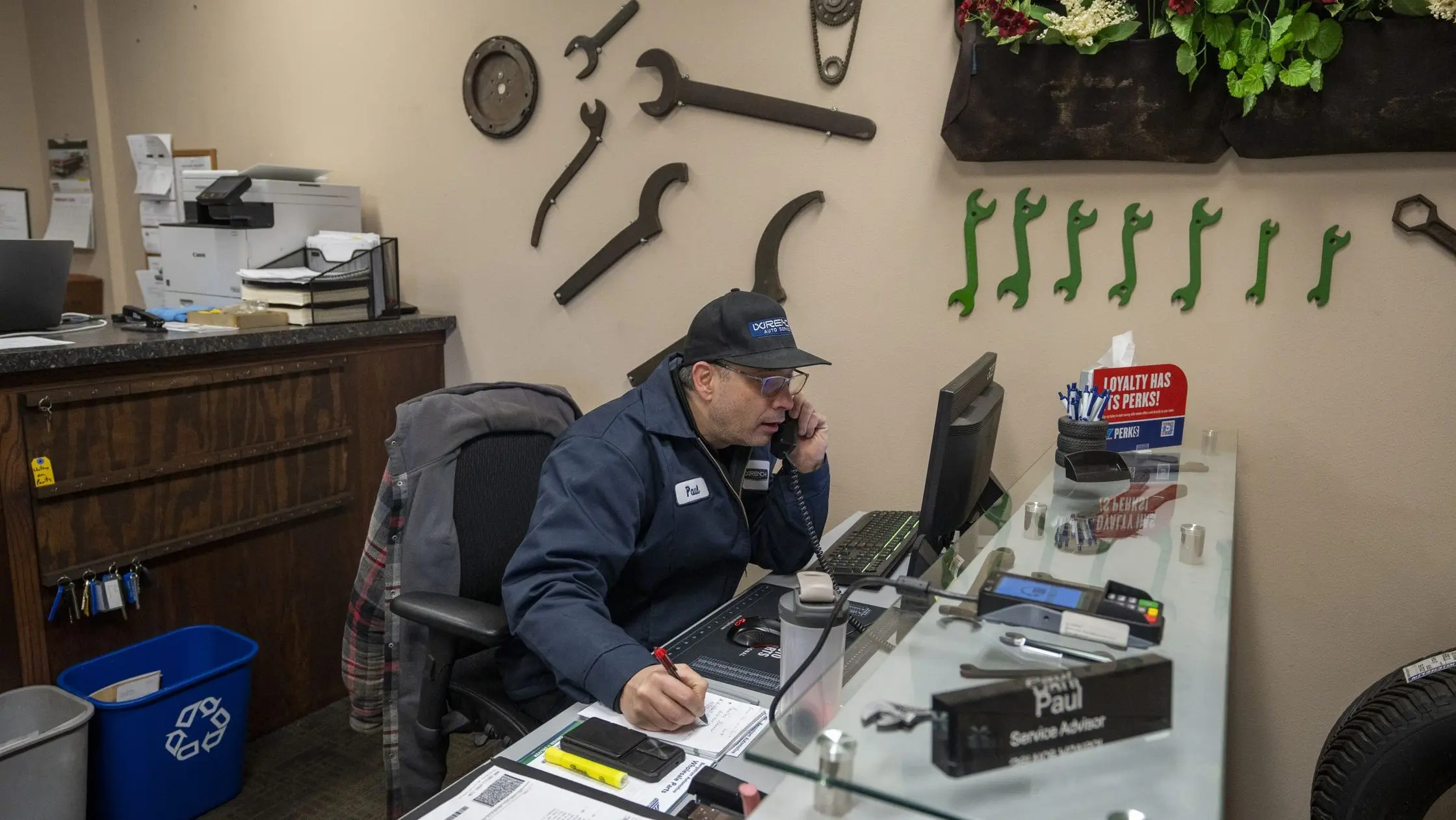 Service advisor Paul at the Wrench Auto Service front desk, on the phone, with wrenches on the wall behind him
