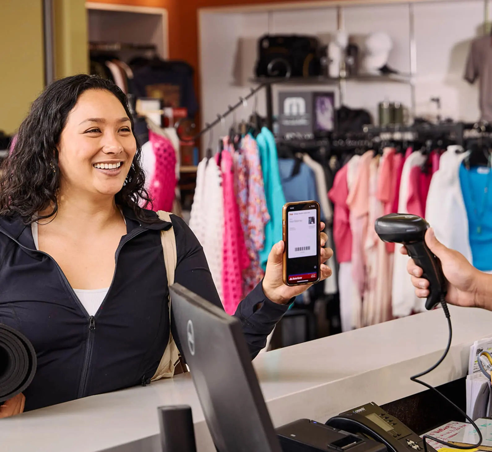 A woman uses her phone to scan a barcode at Village Clubs Camelback clothing store checkout, colorful clothes behind her.